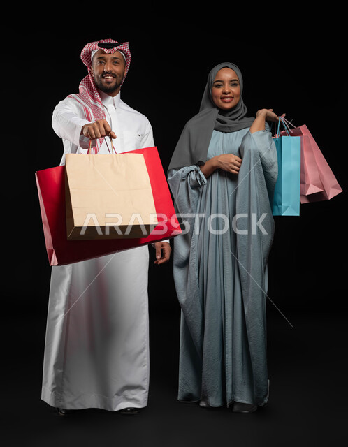Couples participating in choosing purchases, shopping for international and Saudi products, spending a pleasant time buying items on Black Friday, portrait of a Saudi Gulf Arab couple carrying colorful shopping bags, taking advantage of seasonal sales and offers, full length of the body, black background