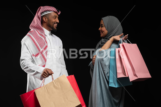 النقاشات والحوارات بين الزوجين، بورتريه لزوجان عربيان خليجيان سعوديان يحملان أكياس التسوق الملونة، الاستمتاع بالشراء وتبضع الأغراض خلال الجمعة البيضاء، استغلال التخفيضات والعروض الموسمية، تسوق المنتجات العالمية والسعودية، خلفية سوداء