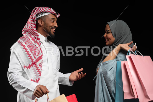الاستمتاع بالشراء وتبضع الأغراض خلال الجمعة البيضاء، النقاشات والحوارات بين الزوجين، بورتريه مقرب لزوجان عربيان خليجيان سعوديان يحملان أكياس التسوق الملونة، استغلال التخفيضات والعروض الموسمية، تسوق المنتجات العالمية والسعودية، خلفية سوداء