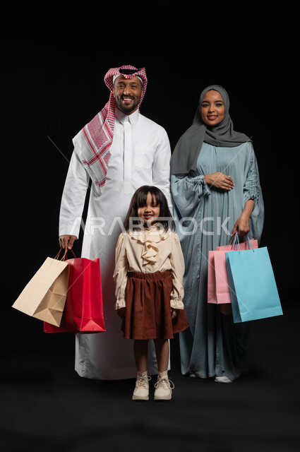 النظر إلى الكاميرا بايماءات البهجة والسرور للقيام بالتسوق العائلي، استغلال التخفيضات والعروض الموسمية، بورتريه لزوجان عربيان خليجيان سعوديان يقفان بإستقامة ويحملان أكياس ملونة برفقة ابنتهم، شراء المنتجات من ماركات عالمية وسعودية، الطول الكامل للجسم، خلفية سوداء