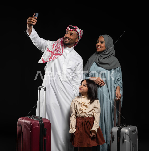 التقاط صور السيلفي التذكارية العائلية، الاستعداد لقضاء إجازة صيفية ممتعة، بورتريه لرجل عربي خليجي سعودي يحمل الهاتف المحمول ويأخذ صورة برفقة عائلته بجانب حقائب السفر، ايماءات السعادة والفرح للذهاب برحلة سياحية، خلفية سوداء