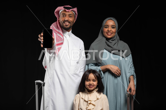 Looking at the camera with gestures of happiness and joy, electronic development and booking a trip via mobile phone, portrait of a Saudi Arabian Gulf man holding a mobile phone with a blank black screen, standing with his family next to travel bags, preparing for a fun summer vacation, black background