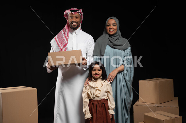 صناديق وحاويات كرتونية مليئة بالأغراض المنزلية، مفهوم الانتقال إلى سكن جديد، بورتريه لعائلة عربية سعودية خليجية تقف باستقامة وتنظر الى الكاميار بتعابير السرور، الاعتناء بالأطفال والاهتمام بهم, قضاء أوقات ممتعة مع الاسرة، خلفيه سوداء