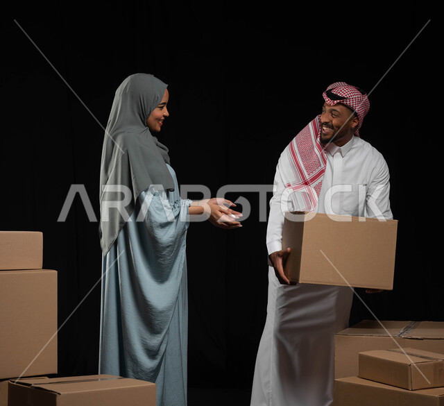تعابير تدل على البهجة والسرور، مفهوم التشارك والتعاون ضمن العائلة السعودية، ترتيب وتنظيم المنزل، بورتريه لزوجين عربيين خليجيين سعوديين يقومان بنقل صناديق الأغراض المنزلية، امتلاك سكن جديد واستقرار الاسرة، خلفية سوداء