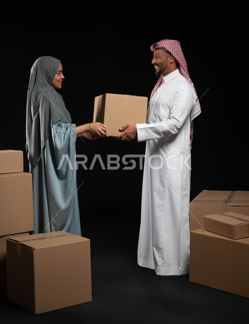 الانتقال الى سكن جديد، التعاون في أداء المهام في العائلة السعودية، تعابير تدل على البهجة والسرور، ترتيب وتنظيم المنزل، بورتريه لزوجين عربيين خليجيين سعوديين يقومان بنقل صناديق الأغراض المنزلية، صورة الجسم بالطول الكامل, خلفية سوداء