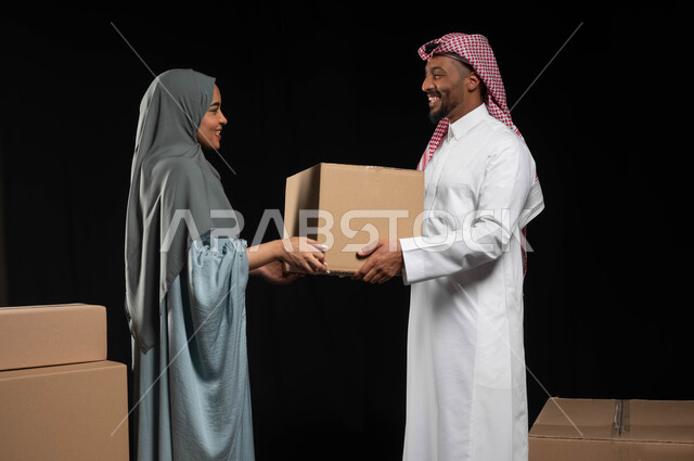 التعاون في أداء المهام في العائلة السعودية، تعابير تدل على البهجة والسرور، ترتيب وتنظيم المنزل، بورتريه لزوجين عربيين خليجيين سعوديين يقومان بنقل صناديق الأغراض المنزلية، امتلاك سكن جديد واستقرار الاسرة، خلفية سوداء