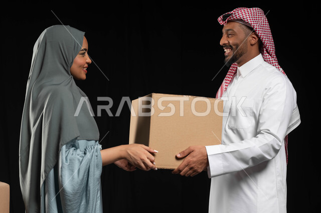إيماءات الفرح والسرور لامتلاك منزل، مفهوم الانتقال الى سكن جديد، التشارك والتعاون ضمن الأسر السعودية، بورتريه مقرب لزوجين عربيين خليجيين سعوديين يتعاونان في ترتيب وتنظيم الأغراض المنزلية، صناديق وحاويات كرتونية، خلفية سوداء