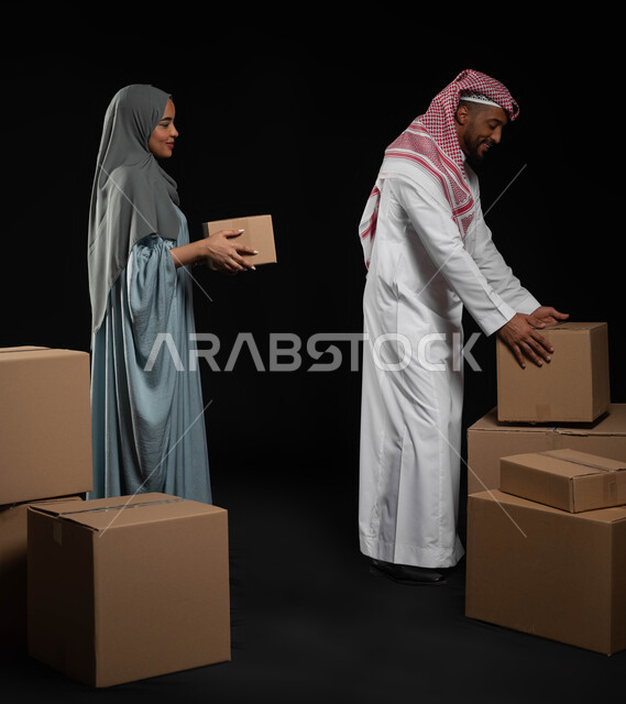 ترتيب وتنظيم المنزل، بورتريه لزوجين عربيين خليجيين سعوديين يقومان بنقل صناديق الأغراض المنزلية، امتلاك سكن جديد واستقرار الاسرة، مفهوم المشاركة والتعاون بحب و سعادة ضمن العائلة السعودية، صورة الجسم بالطول الكامل ,خلفية سوداء