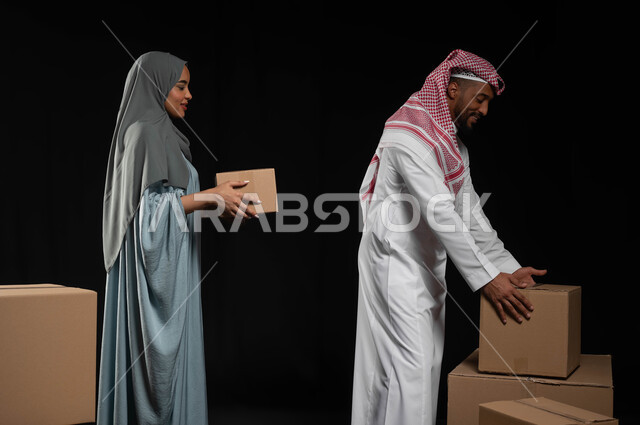 مفهوم المشاركة والتعاون بحب و سعادة ضمن العائلة السعودية، ترتيب وتنظيم المنزل، بورتريه لزوجين عربيين خليجيين سعوديين يقومان بنقل صناديق الأغراض المنزلية، امتلاك سكن جديد واستقرار الاسرة، خلفية سوداء