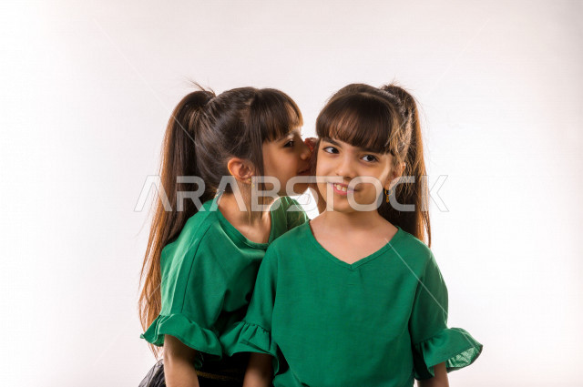 بورتريه لأختان توأم  سعوديتان متشابهتان ، ترتديان ملابس جميلة و متشابهه ، ايماءات وجه واليدين مختلفة ، أطفال توائم ، خلفية بيضاء