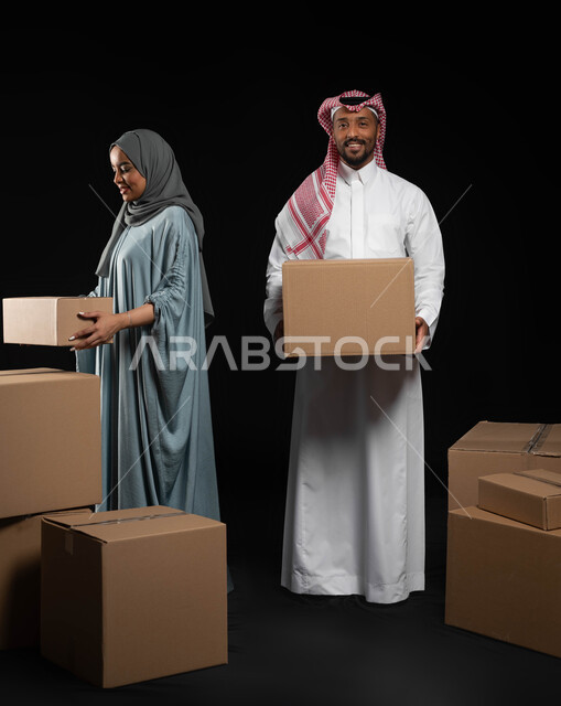 امتلاك سكن جديد واستقرار الاسرة، النظر الى الكاميرا بإيماءات السعادة والسرور، ترتيب وتنظيم المنزل، بورتريه لزوجين عربيين خليجيين سعوديين يقومان بنقل صناديق الأغراض المنزلية، مفهوم التشارك والتعاون ضمن العائلة السعودية، صورة لكامل الجسم, خلفية سوداء