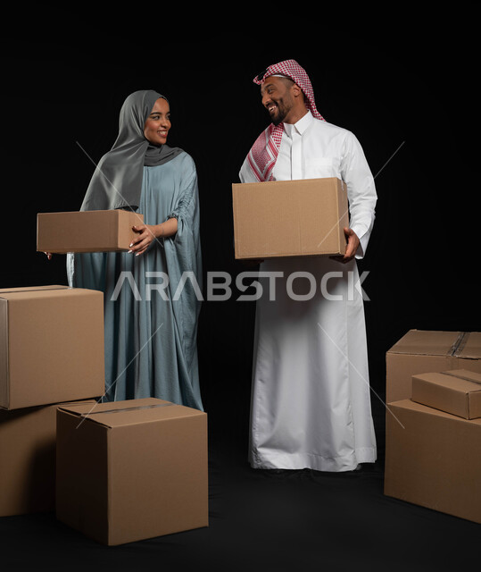 مفهوم المشاركة والتعاون ضمن الأسر السعودية، إيماءات السعادة والسرور لامتلاك سكن جديد، بورتريه لزوجين عربيين خليجيين سعوديين يقومان بترتيب وتنظيم المنزل، صناديق وحاويات كرتونية مليئة بالأغراض المنزلية، صورة الجسم بالكامل, خلفية سوداء