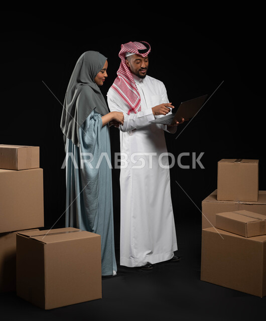 استخدام الوسائل الحديثة في اختيار الأثاث، تعابير البهجة والسرور، مفهوم الانتقال إلى منزل جديد، التعاون والتشارك بين الزوجين في اختيار خطة العمل، بورتريه لرجل عربي خليجي سعودي يرتدي الزي التقليدي يقوم بالنقر على الحاسوب المحمول، صورة لكامل الجسم، خلفيه سوداء
