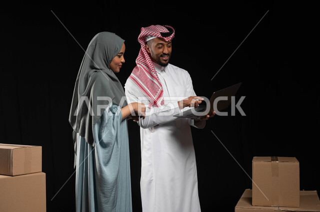 مفهوم الانتقال إلى منزل جديد، استخدام التكنولوجيا الحديثة في أداء المهام والعمل عن بعد، التعاون والتشارك بين الزوجين في اختيار خطة العمل، بورتريه لرجل عربي خليجي سعودي يرتدي الزي التقليدي يقوم بالنقر على الحاسوب المحمول، تعابير البهجة والسرور، خلفيه سوداء