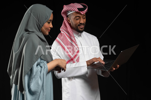 تعابير البهجة والسرور، مفهوم الانتقال إلى منزل جديد، استخدام التكنولوجيا الحديثة في أداء المهام والعمل عن بعد، التعاون والتشارك بين الزوجين في اختيار خطة العمل، بورتريه لرجل عربي خليجي سعودي يرتدي الزي التقليدي يقوم بالنقر على الحاسوب المحمول، خلفيه سوداء