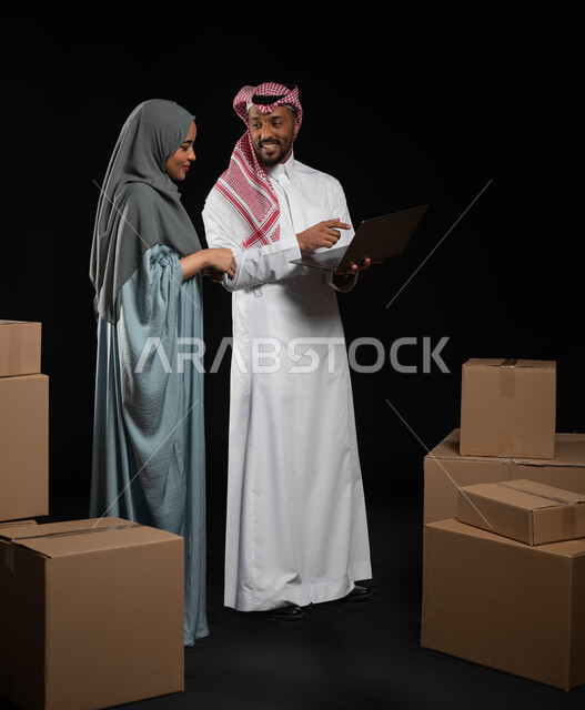 مفهوم الانتقال إلى منزل جديد، استخدام التكنولوجيا الحديثة في أداء المهام، التعاون في اختيار خطة العمل، بورتريه لرجل عربي خليجي سعودي يقوم بالاشارة على الحاسوب المحمول، صناديق وحاويات كرتونية، إيماءات البهجة والسرور، صورة الجسم بالطول الكامل، خلفيه سوداء