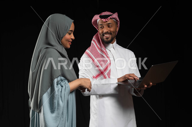 Gestures of joy and happiness, communicating and chatting with family, using modern technology in performing tasks, cooperation and sharing in choosing an action plan, a portrait of a smiling Saudi Gulf Arab couple making a video call via a laptop, black background.