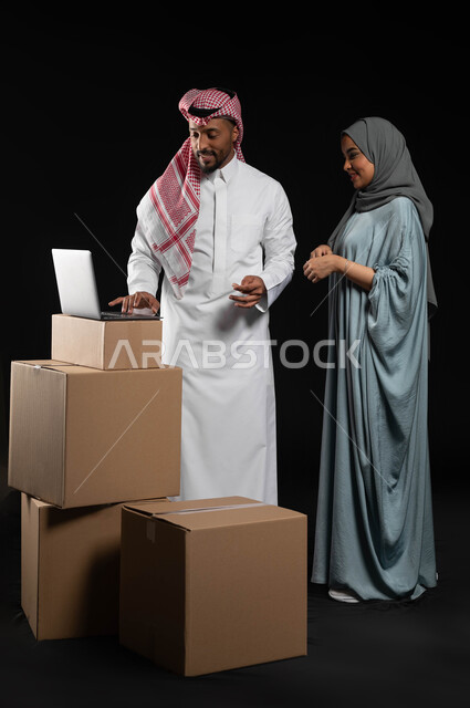 مفهوم الاستقرار العائلي, استخدام الأجهزة التقنية الحديثة، بورتريه لرجل عربي خليجي سعودي يرتدي الزي التقليدي والشماغ برفقة زوجته ينظران الى الحاسوب المحمول، الانتقال الى منزل جديد, كراتين وصناديق جاهزه للتوصيل، صورة الجسم بالكامل, خلفية سوداء