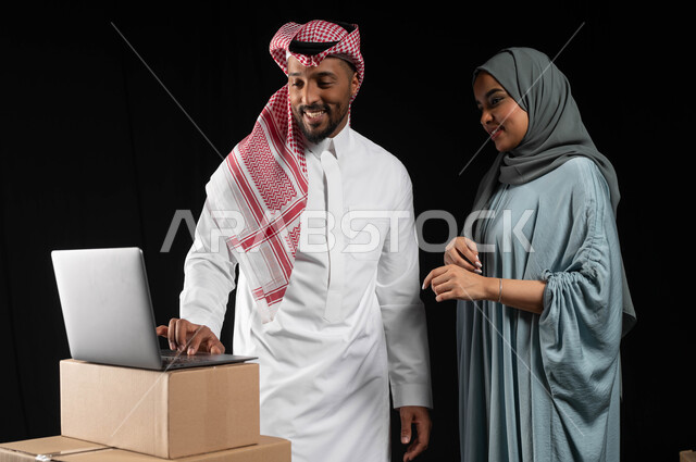 استخدام التكنولوجيا الحديثة في أداء المهام، التعاون في نقل الأغراض المنزليه، بورتريه لرجل عربي خليجي سعودي يقوم بالنقر على الحاسوب المحمول، صناديق وحاويات كرتونية مليئة بالأثاث، مفهوم الانتقال إلى منزل جديد، إيماءات البهجة والسرور، خلفيه سوداء