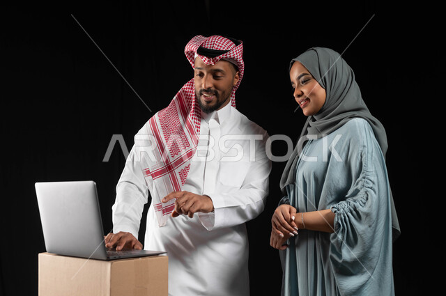 تطور التكنولوجيا وإستخدام الأجهزة التقنية الحديثة، بورتريه لرجل عربي خليجي سعودي يرتدي الزي التقليدي والشماغ برفقة زوجته يشير بيده على الحاسوب المحمول، الانتقال الى منزل جديد, كراتين وصناديق جاهزه للتوصيل، مفهوم الاستقرار العائلي, خلفية سوداء
