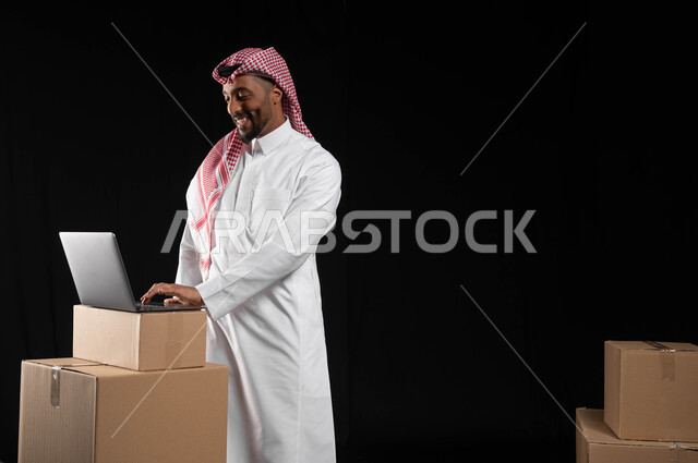 تطور التكنولوجيا وإستخدام الأجهزة التقنية الحديثة، بورتريه لرجل عربي خليجي سعودي مبتسم يرتدي الزي التقليدي والشماغ يقوم بالنظر الى شاشة الحاسوب المحمول بتعابير السرور، صناديق وكراتين جاهزة للتوصيل، خلفيه سوداء