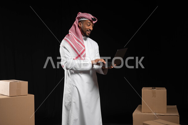 انجاز مهام العمل عن بعد، بورتريه لرجل عربي خليجي سعودي يرتدي الملابس التقليديه يحمل بيده جهاز الحاسوب المحمول بتعابير الدهشة، التواصل و الدردشة مع العملاء ,كراتين وصناديق جاهزة للتوصيل، دمج التقنيات الحديثة المتطورة بالحياة العملية، خلفيه سوداء