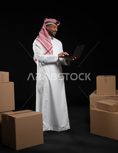دمج التقنيات المتطورة بالحياة العملية، انجاز مهام العمل عن بعد، بورتريه لرجل عربي خليجي سعودي يرتدي الملابس التقليديه يحمل بيده جهاز الحاسوب المحمول، التواصل و الدردشة مع العملاء ,كراتين وصناديق جاهزة للتوصيل، صوره الجسم بالطول الكامل، خلفيه سوداء