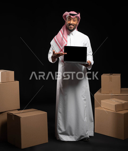 كراتين وصناديق جاهزة للتوصيل، استخدام التقنيات الحديثة ،عرض شاشة سوداء فارغة عبر الحاسوب المحمول، بورتريه مقرب لرجل عربي خليجي سعودي مبتسم يرتدي الملابس التقليديه يشير بيده على اللابتوب، صورة الجسم بالطول الكامل, خلفيه سوداء
