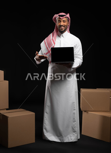 عرض شاشة سوداء فارغة عبر الحاسوب المحمول، بورتريه لرجل عربي خليجي سعودي مبتسم يرتدي الملابس التقليديه يشير بيده على اللابتوب، استخدام التقنيات الحديثة ، كراتين وصناديق جاهزة للتوصيل، صوره الجسم بالطول الكامل، خلفيه سوداء