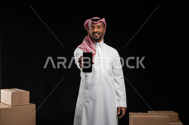 ايماءت الاعجاب باستخدام التقنيه والتكنولوجيا الحديثه، بورتريه لرجل عربي خليجي سعودي مبتسم يرتدي الثوب التقليدييحمل بيده الجوال بشاشة سوداء فارغة، كراتين وصناديق جاهزة للتوصيل، التواصل مع شركات نقل الاثاث، خلفيه سوداء