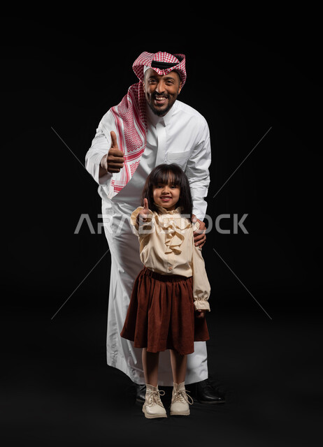 تقوية الروابط والعلاقات مع الأطفال بالعطف و الحنان، مفهوم الترابط الأسري بين أفراد العائلة، بورتريه لرجل عربي خليجي سعودي يرتدي الملابس التقليديه يقف خلف ابنته ويرفعان اصابع الابهام للاعلى بتعابير علامة الجودة، صورة الجسم بالكامل, خلفيه سوداء