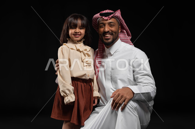 الشعور بالأمان والحب مع الآباء، بورتريه لأب وأبنته عربيان خليجيان سعوديان مبتسمان ينظران إلى الكاميرا بإيماءات البهجة والسرور، مفهوم الإعتناء بالأبناء والإهتمام بهم، العلاقات الأسرية الناجحة، خلفية سوداء
