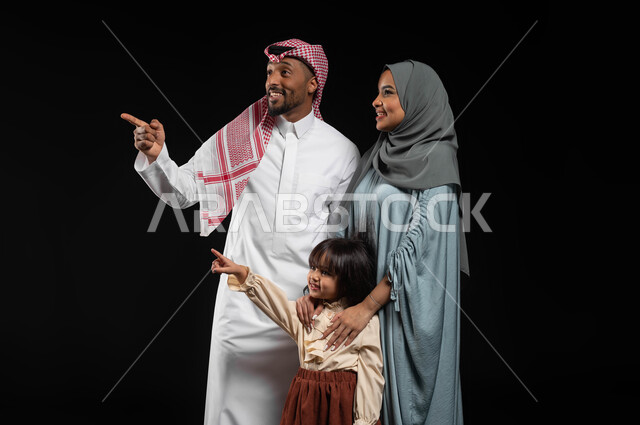 The concept of raising children on the right path, a portrait of a Saudi Gulf Arab family looking and pointing with their index finger in order to attract attention, gestures of happiness and pleasure at seeing something, future goals and ambitions, a family atmosphere full of love, a black background