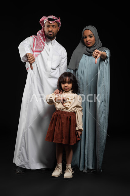 الإشارة بإصبع الإبهام الى الأسفل والإنزعاج من شيء ما, ايماءات وجه ويدين تدل على الرفض وعدم الموافقة, بورتريه لعائلة عربية خليجية سعودية تقف بإستقامة وتقوم بالنظر إلى الكاميرا, صورة الجسم بالطول الكامل, خلفية سوداء