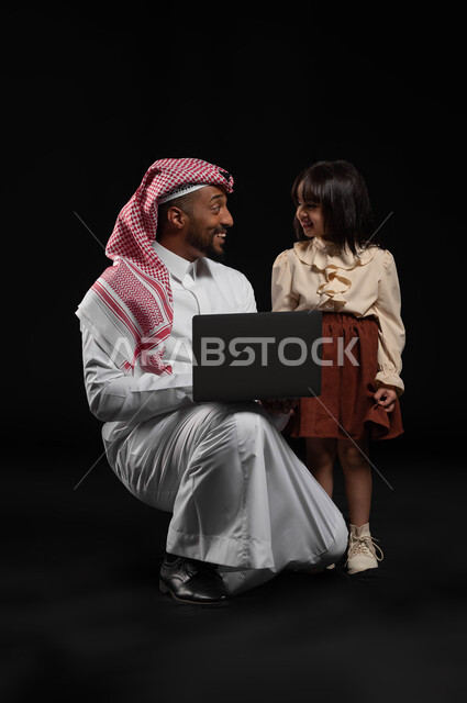 قضاء الأوقات السعيدة مع الأهل, مفهوم الرعاية والإهتمام بالأبناء, بورتريه لرجل عربي خليجي سعودي يرتدي الثوب التقليدي والشماغ يحمل لابتوب برفقة ابنته, إجراء مكالمات الفيديو والبثوث المباشرة عبر الإنترنت, متابعة الحصص الدراسية أونلاين, خلفية سوداء