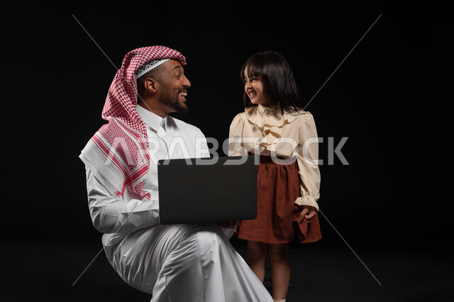 Sharing happy times with parents, the concept of care and concern for children, a portrait of a Saudi Gulf Arab man wearing a traditional thobe and shemagh carrying a laptop with his daughter, making video calls and live broadcasts over the Internet, following classes online, black background