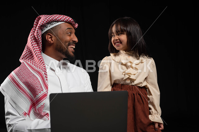 إجراء مكالمات الفيديو والبثوث المباشرة عبر الإنترنت, متابعة الحصص الدراسية أونلاين, مفهوم الرعاية والإهتمام بالأبناء, بورتريه مقرب لرجل عربي خليجي سعودي يرتدي الثوب التقليدي والشماغ يحمل لابتوب برفقة ابنته, مشاركة الأوقات السعيدة مع الأهل, خلفية سوداء
