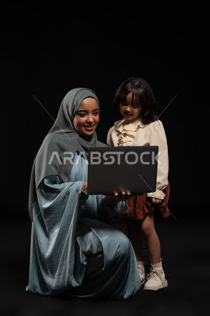 متابعة الحصص الدراسية أونلاين, مفهوم الرعاية والإهتمام بالأبناء, بورتريه لإمرأة عربية خليجية سعودية محجبة ترتدي عباءة وتحمل لابتوب برفقة ابنتها, إجراء مكالمات الفيديو والبثوث المباشرة عبر الإنترنت, مشاركة الأوقات السعيدة مع الأهل, خلفية سوداء