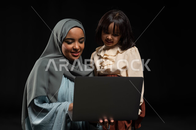 مفهوم الرعاية والإهتمام بالأبناء, بورتريه مقرب لإمرأة عربية خليجية سعودية محجبة ترتدي عباءة وتحمل لابتوب برفقة ابنتها, متابعة الحصص الدراسية أونلاين, إجراء مكالمات الفيديو والبثوث المباشرة عبر الإنترنت, مشاركة الأوقات السعيدة مع الأهل, خلفية سوداء
