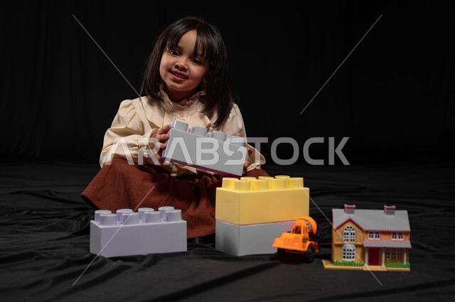 الأنشطة الترفيهية والتعليمية للأطفال، بورتريه لطفلة عربية خليجية سعودية ترتدي الزي الكاجوال تجلس وتقوم بتركيب المكعبات، تعزيز المهارات الذهنية والفكرية لدى الأطفال، إيماءات الفرح والسعادة لقضاء أوقات ممتعة باللعب واللهو، خلفية سوداء
