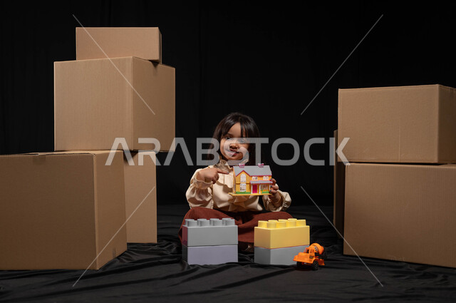Moving to a new home, recreational and educational activities for children, portrait of a Saudi Arabian Gulf girl wearing a casual outfit sitting and holding in her hand a toy in the shape of a small house with cardboard boxes next to it, gestures of joy and happiness to enjoy playing and assembling cubes, black background