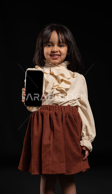 Using modern and advanced technology and techniques, a portrait of a smiling Saudi Gulf Arab girl standing upright and holding a mobile phone with a blank black screen, electronic marketing and displaying mobile services and applications, looking at the camera with gestures of happiness and pleasure, black background.