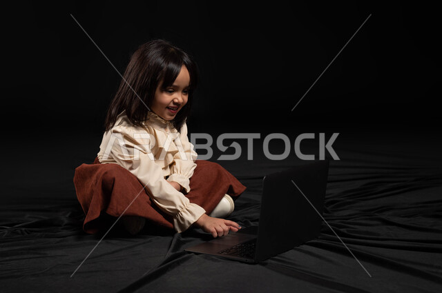 مهارة و احتراف وظائف الحاسوب و تكنولوجيا المعلومات منذ الصغر،دور التقنية الحديثة في توفير راحة أكبر للأفراد، بورتريه لفتاة عربية خليجية سعودية مبتسمة ترتدي الزي الكاجوال تنظر الحاسوب المحمول بتعابير السرور و تقوم بالنقر على لوحة المفاتيح، خلفيه سوداء