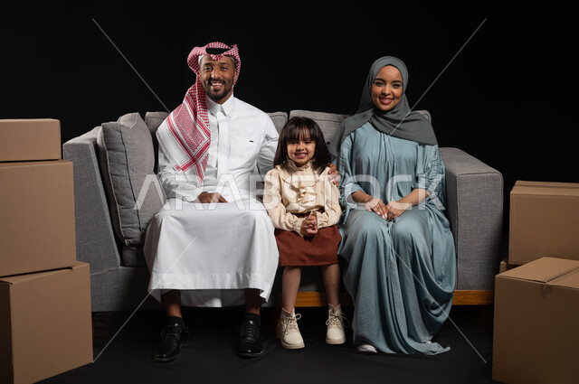 Expressions of joy at owning a new home, the concept of family stability, a mother’s care and attention to her children, the concept of a happy family, a portrait of a Saudi Gulf Arab family sitting on the sofa and looking at the camera with happy gestures, cardboard containers and boxes, black background