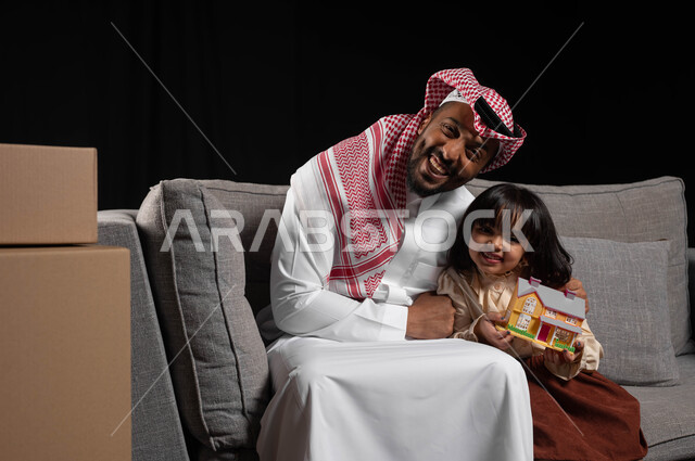 النظر الى الكاميرا بايماءات السعاده لامتلاك سكن جديد، أهمية مشاركة الوقت الترفيهي مع الأبناء, بورتريه لأب عربي خليجي سعودي يرتدي الملابس التقليديه يجلس على الاريكه يقوم بعانقة ابنته الصغيره تحمل بيدها مجسم على شكل منزل, حاويات وصناديق كرتونيه، خلفيه سوداء