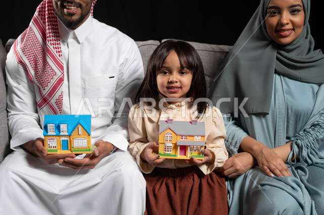 الإنتقال الى منزل جديد، بورتريه لعائلة عربية خليجية سعودية تجلس على الاريكه وتنظر الى الكاميرا بتعابير السعادة ,حمل مجسم صغير على شكل منزل، حاويات وصناديق كرتونيه، مفهوم الاستقرار العائلي, خلفيه سوداء