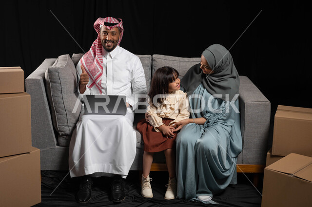 إجراء الأب لمكالمات الفيديو والبثوث المباشره من خلال الحاسب المحمول، بورتريه مقرب لعائله عربيه خليجيه سعوديه تجلس على الأريكه، الاستمتاع بالحديث بين الام والبنت، صناديق وحاويات كرتونية، خلفيه سوداء