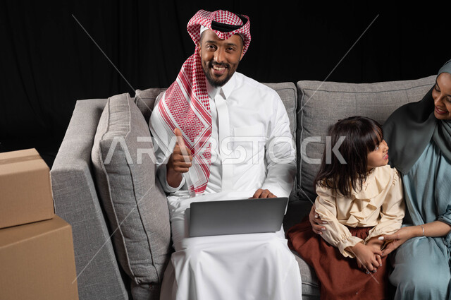 تبادل الاحاديث بين الأم و ابنتها، بورتريه لرجل عربي خليجي سعودي يرتدي الزي التقليدي يحمل الحاسوب المحمول يرفع اصبعه بعلامه الجودة وينظر الى الكاميرا بتعابير السرور، مفهوم الاستقرار العائلي، صناديق وحاويات كرتونية، الاستمتاع بالسكن الجديد، خلفيه سوداء