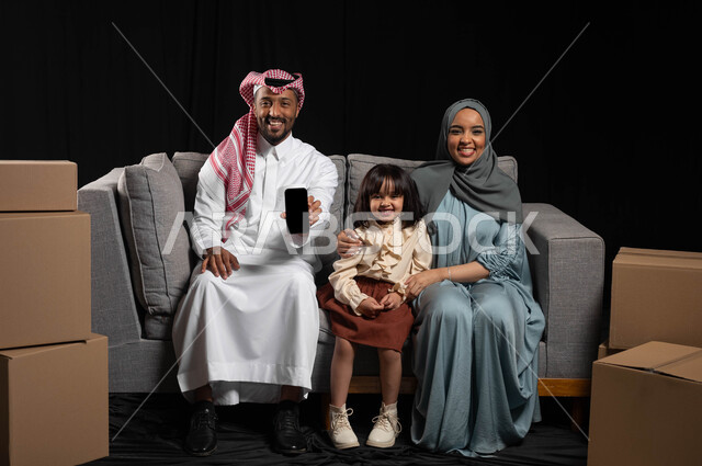 صناديق وحاويات كرتونية، مفهوم الاستقرار العائلي، عرض شاشة سوداء فارغه عبر الجوال، استخدام تطبيقات الهاتف المحمول للبحث عن شركات نقل الاثاث للمنزل الجديد، بورتريه لعائلة عربية خليجية سعودية تجلس على الاريكه وتنظر الى الكاميرا بإيماءات السعادة، خلفيه سوداء