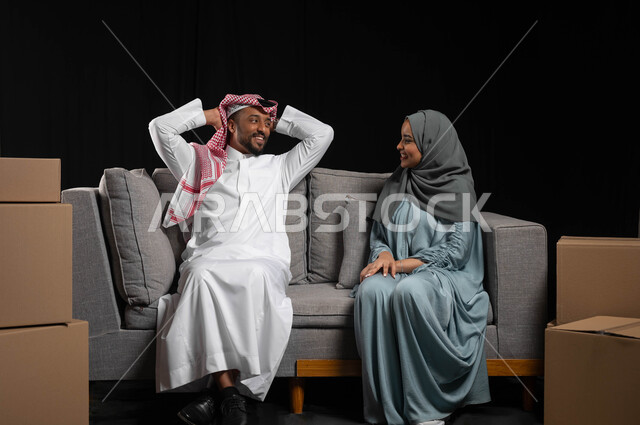 الانتقال إلى سكن جديد، الترابط الأسري والحفاظ على الاستقرار العائلي، بورتريه لرجل عربي خليجي سعودي مبتسم يرتدي الزي التقليدي يجلس على الاريكة الرمادية و يضع يديه وراء رأسه بايماءات تبادل الاحاديث مع الزوجة ,صناديق وحاويات كرتونية، خلفيه سوداء